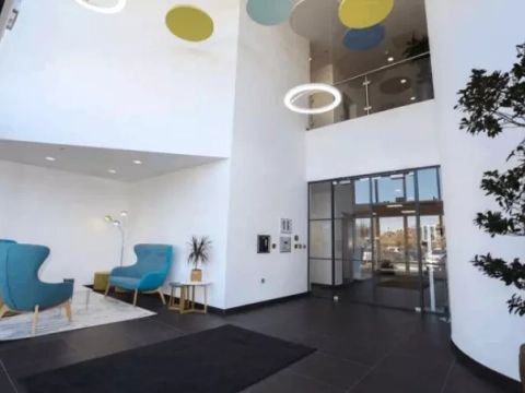 Modern office lobby with blue chairs, white walls, colorful ceiling accents, and large windows. A plant is near the entrance, and a second level is visible with glass railings.