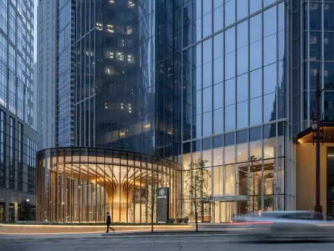 Modern glass skyscraper with a curved entrance and illuminated interior. A car passes by on the street in front of the building.